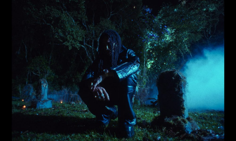 A imagem é um corte de um videoclipe do cantor Matuê e mostra uma figura masculina agachada em um cemitério à noite, cercada por árvores e lápides, envolta por fumaça azulada. A cena evoca luto, introspecção e ritual, transformando o espaço da morte em território de contemplação e memória.