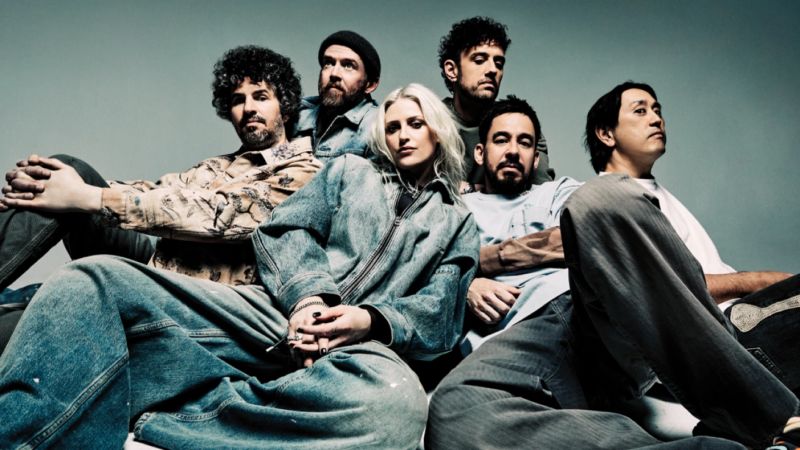 Seis integrantes da banda Linkin Park sentados juntos em estúdio, vistos de baixo para cima, vestindo roupas largas em tons neutros e jeans. Ao centro, uma mulher com cabelo platinado cruza as pernas e encara a câmera, enquanto os demais membros ao redor mantêm posturas relaxadas e expressões sérias ou contemplativas.