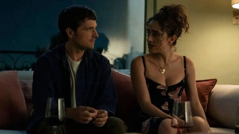 Cena da série I Love LA. Na imagem, há um homem branco de cabelos castanhos com uma blusa branca e uma jaqueta azul escuro. Ele está olhando para uma mulher branca de cabelos ondulados que usa um vestido preto com estampas de flores e um colar dourado de coração. Os dois se encaram e estão sentados em um sofá. Na frente dos dois, há duas taças com vinho dentro.