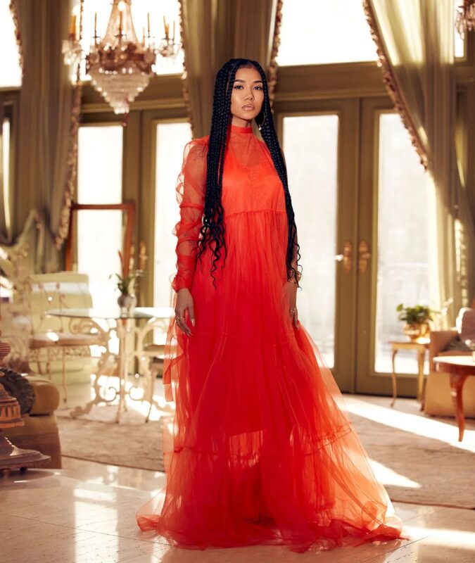 Na imagem, há uma mulher com longas tranças escuras usando um vestido longo vermelho-alaranjado com mangas longas. Ela está em um salão elegante com grandes janelas, cortinas luxuosas e um lustre de cristal ao fundo. A luz natural ilumina o ambiente, realçando a textura translúcida do vestido.