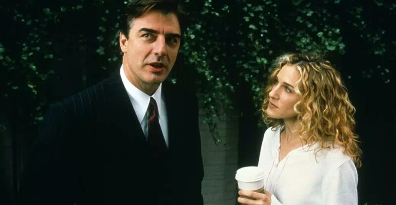 Imagem de uma cena da série Sex and the City. Foto tirada em uma calçada, ao fundo aparece uma coluna branca que faz parte de uma casa e vários galhos pendurados do topo até a metade da foto. No lado esquerdo, Mr. Big, um homem branco de cabelos curtos marrom, usando um terno preto com linhas quase não aparentes da mesma cor, uma camisa social branca e uma gravata vinho. Na direita, Carrie, de perfil, uma mulher branca, de olhos claros e cabelos dourados cacheados, usando uma jaqueta branca e segurando um café. 
