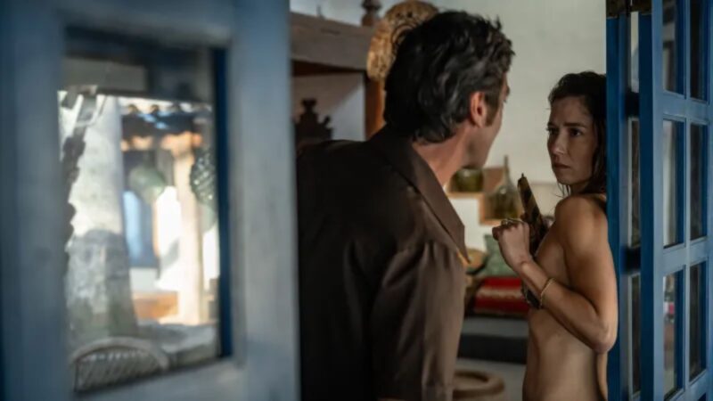 Cena de Ângela Diniz: Assassinada e condenada. Ângela, uma mulher de pele clara está próxima a uma porta azul. Ela tem cabelo castanho escuro, veste um biquíni e apresenta uma expressão tensa, olhando para um homem à sua frente. Doca, de costas para a câmera, tem cabelo escuro e veste uma camisa marrom escuro. O cenário inclui alguns móveis e objetos ao fundo.