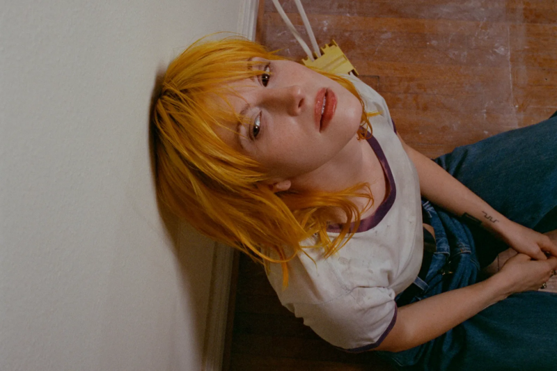 Texto Alternativo: Fotografia de Hayley Williams. Na imagem capturada de cima para baixo, a cantora está sentada em um chão de madeira e com cabeça encostada na parede, direcionada para cima, mas seu olhar encara seu lado direito. Williams tem cabelo amarelado e a boca um pouco aberta, usa uma calça jeans e uma camiseta branca com gola magenta. O chão está manchado de poeira.