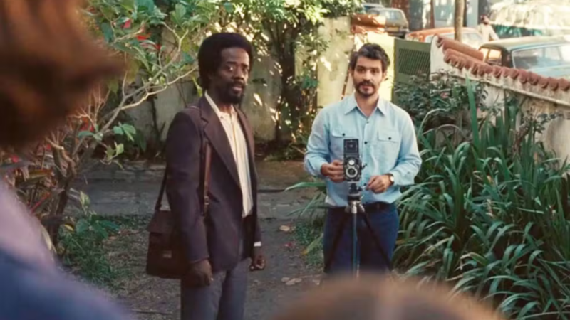 Philipp Lavra, à direita, sorri levemente enquanto manuseia uma câmera fotográfica antiga de médio formato sobre um tripé. Ele veste camisa azul clara. Ao seu lado, um homem negro de terno marrom observa. O cenário é um jardim ensolarado com muro branco e plantas verdes.