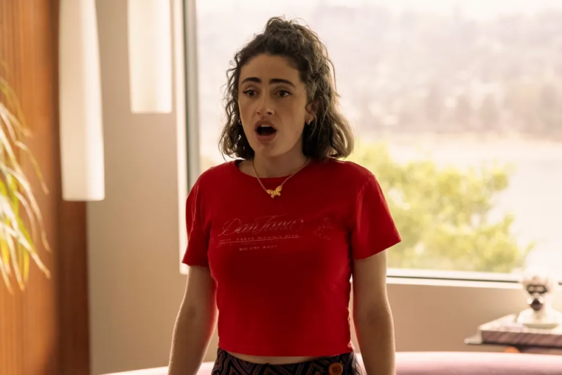 Cena da série I Love LA. Na imagem, há uma mulher branca de cabelos ondulados que veste uma blusa vermelha com um colar dourado de borboleta. Ela está em pé e com uma expressão de espanto. 