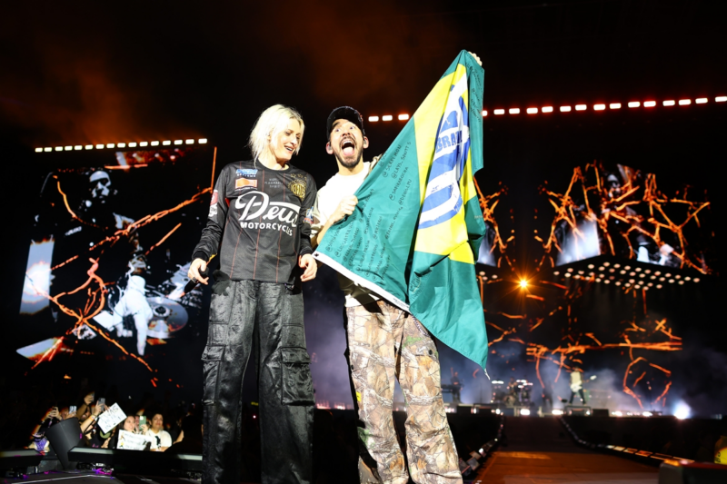 Uma mulher com cabelo platinado e um homem com boné sobre o palco durante um show, sorrindo e segurando juntos uma bandeira do Brasil aberta. Ao fundo, telões exibem imagens em preto e branco, enquanto luzes quentes e a plateia lotada reforçam o clima de celebração e conexão com o público.