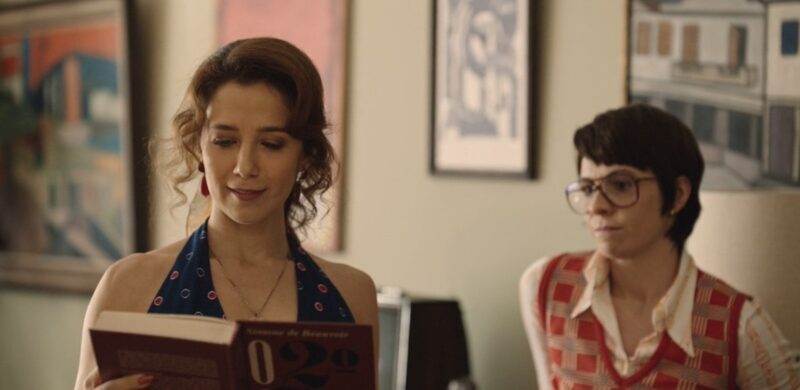 Cena de Ângela Diniz: Assassinada e Condenada. Duas mulheres estão em um ambiente interno bem iluminado, com quadros pendurados na parede ao fundo. À esquerda, Ângela, uma mulher de pele clara e cabelo castanho ondulado preso lê um livro, olhando para baixo. Ela usa um vestido azul-marinho sem mangas. À direita, um pouco mais ao fundo, outra mulher de pele clara e cabelos castanhos curtos observa a leitura. Ela usa óculos e veste uma camisa clara com um colete em tons de vermelho e laranja. No fundo, aparece uma parede branca com quadros pendurados. 