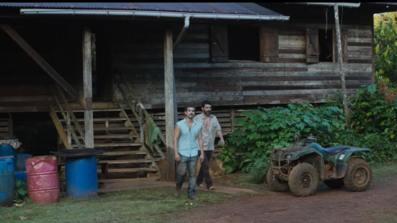 Plano aberto mostra uma casa de madeira desgastada sobre palafitas. No terreiro de terra batida à frente da casa, Philipp Lavra e Rômulo Braga caminham lado a lado. Há um quadriciclo verde estacionado à direita e barris azuis e rosados à esquerda.