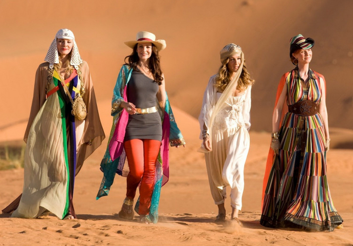 Imagem da cena do filme Sex and the City 2. Quatro mulheres andando lado a lado em um deserto, vestidas com roupas coloridas que misturam elementos ocidentais e orientais. Da esquerda para a direita: Samantha usa um vestido longo bege com detalhes coloridos e um acessório na cabeça; Charlotte veste calça vermelha, blusa/vestido cinza e chapéu de palha, com um kimono colorido esvoaçante; Carrie usa um conjunto branco com faixa na cabeça; e Miranda está com um vestido longo de listras coloridas e chapéu preto com faixa. 
