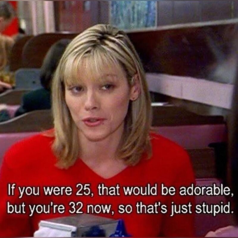 Imagem da série Sex and the City, mostra Samantha, uma mulher loira com cabelo liso na altura dos ombros, usando uma blusa vermelha, sentada em uma cabine de restaurante ou lanchonete. Ela está em primeiro plano, ligeiramente voltada para alguém fora de quadro, com uma expressão de leve ironia ou reprovação. Atrás dela, há outras pessoas sentadas. Na parte inferior da imagem há uma legenda em inglês que diz: "If you were 25, that would be adorable, but you're 32 now, so that's just stupid."