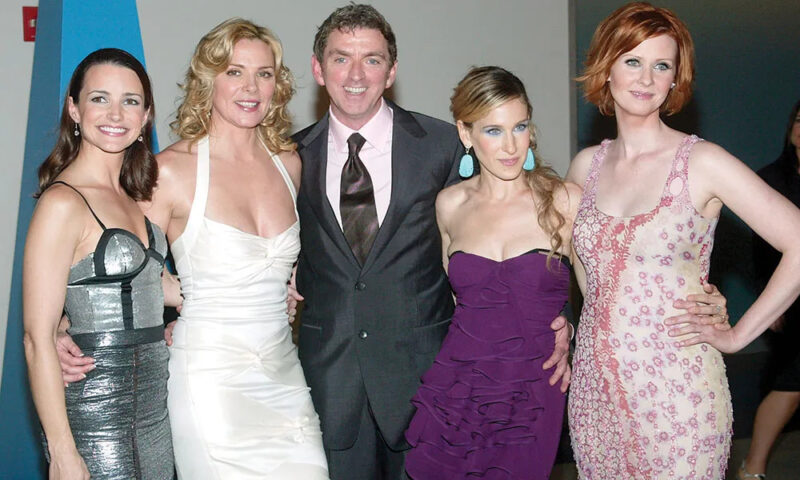 Fotografia de Michael Patrick King com as quatro atrizes principais de Sex and the City. À esquerda, Kristin Davis, uma mulher de peles claras, cabelo marrom na altura do ombro, usando um vestido de alças finas prateado com detalhes pretos. Ao seu lado, Kim Cattrall, uma mulher de pele clara, cabelo loiro ondulado, usando um vestido branco de alças grossas, com as mãos na cintura das pessoas ao seu lado. No meio da fotografia, Michael Patrick King, um homem branco de cabelo curto e escuro, olhos azuis, usando terno e calça social pretos, com uma camisa social branca por baixo e uma gravata preta. Ao seu lado, Sarah Jessica Parker, uma mulher branca, loira com cabelos presos, de olhos azuis, usando um vestido tomara que caia roxo com babados e um brinco verde água. Por fim, Cynthia Nixon, uma mulher branca, ruiva de cabelos curtos, de olhos claros, usando um vestido bege com flores rosas. 