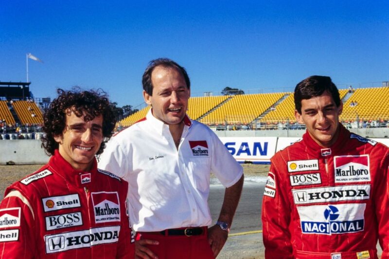 A cena mostra três homens sorridentes posando juntos em um autódromo ensolarado, dois deles vestem macacões de corrida vermelhos repletos de logotipos de patrocinadores, enquanto o terceiro entre os dois pilotos, ao centro, usa camisa branca da equipe com detalhes em vermelho Marlboro e uma calça da mesma cor junto a um cinto preto. Ao fundo, uma arquibancada amarela quase vazia cria o clima de preparação para um grande evento automobilístico.
