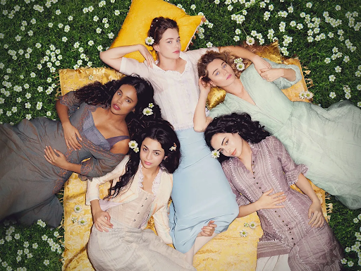  Imagem promocional da série The Buccaneers. Cinco mulheres estão deitadas de costas em um campo verde cheio de pequenas flores brancas. Elas usam vestidos de cores suaves e românticas, com flores presas no cabelo. Elas estão dispostas em círculo com as cabeças próximas e olhando para cima