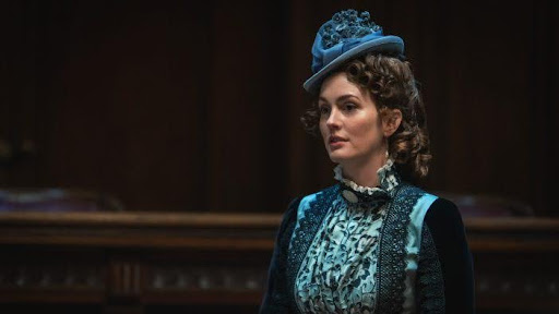 Cena da série The Buccaneers. Vemos Nell (Leighton Meester), uma mulher branca com cabelo ruivo, preso em cachos e enrolado ao redor da cabeça. Ela usa um chapéu azul decorado com flores e um vestido azul claro com detalhes em renda preta, gola alta com babados brancos. O fundo está desfocado e escuro