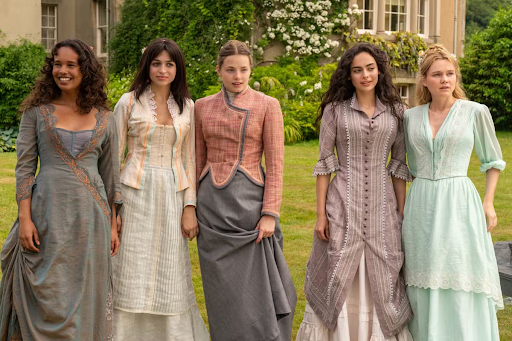 Cena da série The Buccaneers. Cinco mulheres estão em pé, lado a lado, segurando suas saias ou mãos, olhando para a direita. Elas vestem roupas de época em tons pastel e terrosos, com detalhes bordados ou rendados. O jardim atrás delas tem plantas verdes e uma casa antiga parcialmente visível 