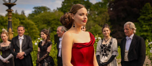Cena da série The Buccaneers. Vemos na imagem Nan, uma mulher branca, de cabelo preso para trás, usando um vestido vermelho elegante e tomara que caia. Ela está de perfil para a direita e tem um batom vermelho. Ao fundo, há um grupo de pessoas vestidas formalmente em trajes de época, em um jardim com vegetação verde 