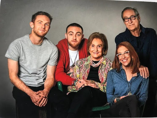 Fotografia de estúdio que mostra Mac Miller com sua família, posando juntos diante de um fundo neutro. Ao centro da imagem, sentada, está uma senhora idosa de expressão gentil, vestindo uma jaqueta estampada colorida com detalhes em rosa, verde e dourado, colares de pérolas e uma blusa preta. Ela segura as mãos de Mac Miller, que está logo atrás, à esquerda. Mac Miller aparece sentado parcialmente atrás dela, com as mãos cruzadas sobre os joelhos. Ele usa uma camiseta branca, uma corrente prateada e um casaco vermelho. À esquerda dele está um homem jovem de camiseta cinza e calça preta, com os braços apoiados nas pernas. À direita da senhora, também sentada, está uma mulher ruiva de óculos, blusa azul e expressão sorridente. Em pé atrás dela está um homem mais velho, de óculos, cabelos grisalhos e camisa azul-escura, sorrindo levemente com uma mão apoiada no ombro da mulher à sua frente. 