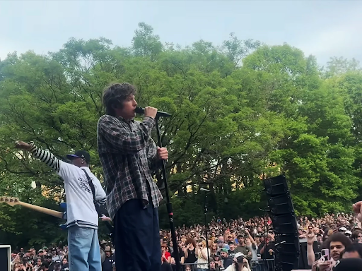Cantor se apresenta em um palco ao ar livre, em um lugar arborizado, segurando um microfone em frente a uma grande multidão. Ele é um homem jovem, branco, de cabelos cacheados, veste camisa xadrez e calça azul, ele canta enquanto um guitarrista ao fundo aponta para o alto. Há uma multidão ao redor.