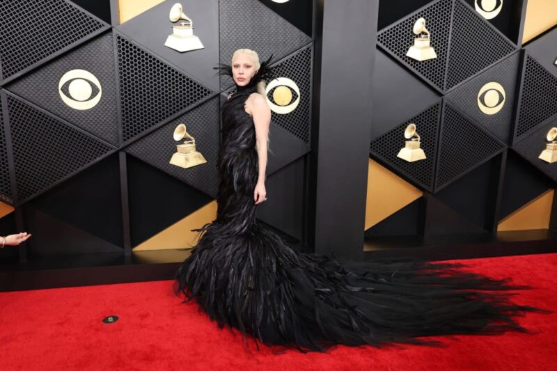 Fotografia de corpo inteiro de Lady Gaga no tapete vermelho do Grammy Awards. A artista está posicionada de perfil para a câmera, olhando por cima do ombro com uma expressão séria. Ela usa um vestido longo preto composto inteiramente por penas sintéticas e tule, que criam uma textura volumosa. O vestido possui uma gola alta e detalhes que sobem como asas atrás do pescoço. A saia se estende em uma longa cauda dramática que se espalha pelo tapete vermelho. Lady Gaga está com o cabelo loiro platinado e uma maquiagem suave. O fundo apresenta o painel oficial do evento em preto e dourado com o logotipo da CBS e o símbolo do gramofone.