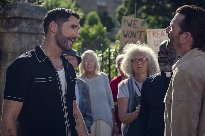 Cena do filme O Clube do Crime das Quintas-Feiras.Na imagem, está Jason (Tom Elis), homem branco, de barba e cabelos curtos e escuros, vestindo uma blusa preta sobre uma camiseta branca aparece discutindo com Ian Ventham, que está de perfil. Ele é um homem branco de cabelos castanhos e veste uma jaqueta bege. Ao fundo, estão moradores de Coopers Chase segurando cartazes durante um protesto. 