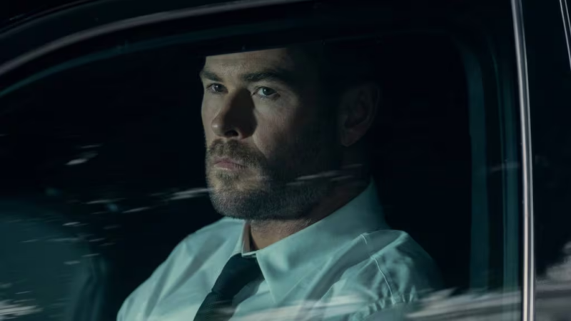 Cena de Caminhos do Crime. Close-up do ator Chris Hemsworth dentro de um carro à noite. Ele tem uma expressão séria e pensativa, olhando para o lado. Está vestindo uma camisa social branca e uma gravata preta. O reflexo das luzes da cidade e borrões de movimento aparecem no vidro da janela em primeiro plano, criando uma atmosfera de suspense ou drama.