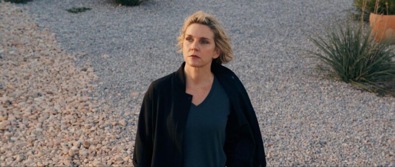 Rhea Seehorn, mulher branca, loira e de olhos azuis, está posicionada no centro da imagem. Ela veste uma camisa azul escura com um casaco preto aberto por cima. A foto foi tirada em um ângulo que destaca o chão de pedras como fundo principal. No canto superior direito, aparecem algumas plantas.