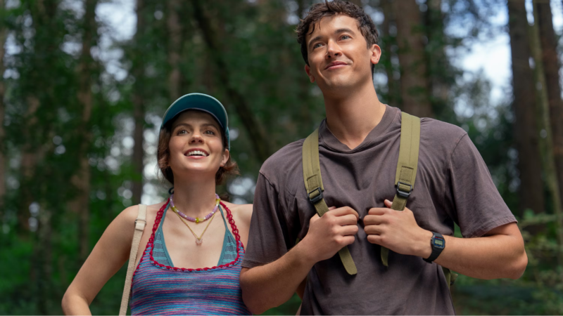 Em uma floresta, Poppy e Alex estão no centro da imagem, com uma expressão de felicidade olhando para algo. Ela está com uma regata e biquíni listrado e um boné azul, enquanto ele está com uma mochila nas costas verde e uma camiseta cinza-escuro.