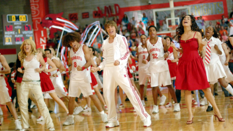 Cena do filme High School Musical. Na imagem, o elenco principal do filme está no ginásio da escola East High. Eles estão performando a faixa We’re All in This Together e vestem roupas como calças e regatas brancas, vestidos e roupas vermelhas. Da esquerda para a direita, há uma mulher branca de cabelo louro e ondulado, um homem negro de cabelo cacheado e castanho claro, um homem branco de cabelo liso cor de mel, um homem negro de cabelo crespo castanho escuro e uma mulher branca de cabelo castanho escuro ondulado.