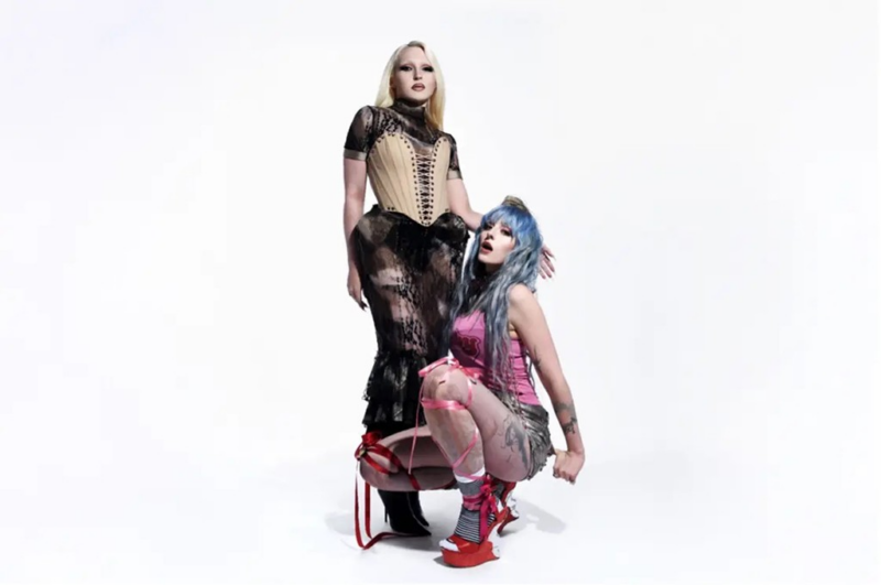 Em um fundo branco minimalista, Ashnikko e Cobrah posam juntas. Cobrah é uma mulher branca de cabelos loiros lisos e está em pé, usando um vestido preto transparente de renda com um espartilho bege estruturado. Ao lado dela, Ashnikko está agachada usando uma regata rosa com um shorts e sapatos vermelhos de plataforma, fitas rosas e vermelhas entrelaçam em suas pernas