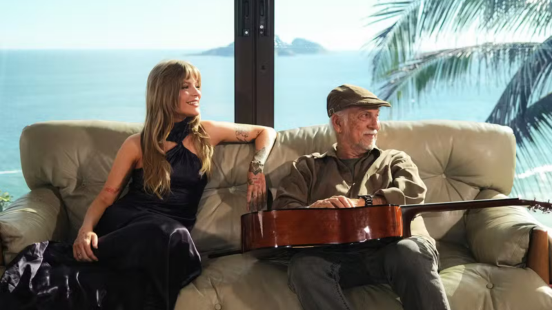 Luísa Sonza e Roberto Menescal sentados confortavelmente em um sofá de couro bege claro. Eles estão em uma sala com uma grande janela de vidro ao fundo, que oferece vista para o mar e ilhas. Luísa, à esquerda, usa o vestido roxo da capa e sorri olhando para a frente. Roberto Menescal, à direita, veste uma camisa marrom e uma boina; ele segura um violão de madeira no colo e olha para a paisagem externa com um leve sorriso.