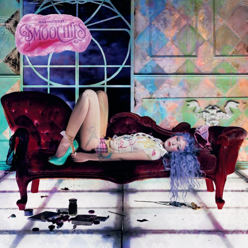 Ashnikko aparece uma pessoa deitada de costas em um sofá vermelho de veludo, em um cenário interno colorido e onírico. Ela tem cabelos longos azuis, usa meia-calça clara, short xadrez, camiseta clara e stampada e salto verde. No chão branco, há pequenos objetos espalhados. Ao fundo, paredes com padrões geométricos e cores iridescentes; no canto superior esquerdo aparece a palavra Smoochies em uma forma rosa semelhante a um chiclete.