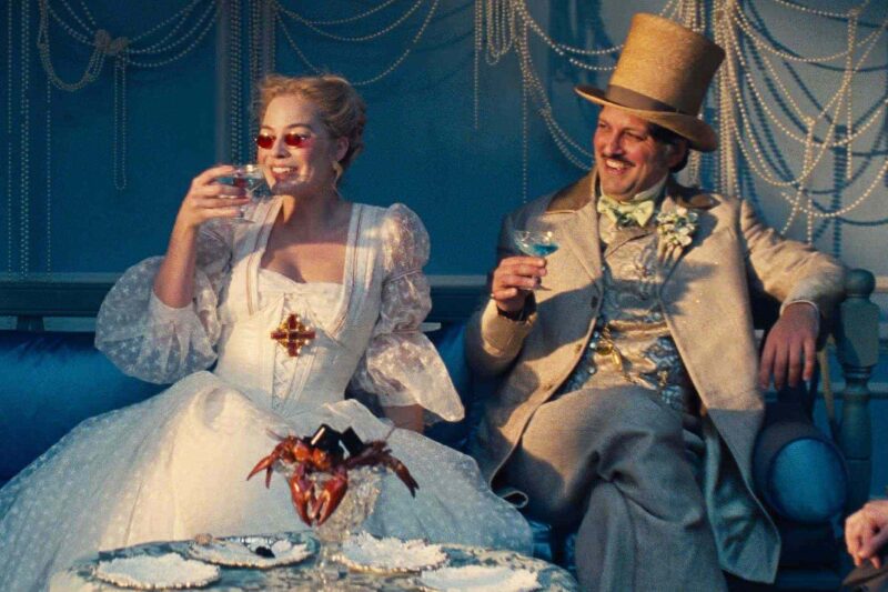 Cena de “O Morro dos Ventos Uivantes”. À esquerda, Catherine, uma mulher de pele clara e cabelo loiro preso usa vestido branco volumoso com mangas bufantes e óculos de lentes vermelhas. À direita, um homem de pele mais escura, cabelo e bigode castanhos veste terno bege com detalhes dourados, colete e cartola. Eles seguram taças que contém um líquido azul. Ao fundo, a decoração é composta por fios de pérolas sobre parede azul. Na mesa à frente deles, há uma espécie de caranguejo servido em um recipiente de vidro.