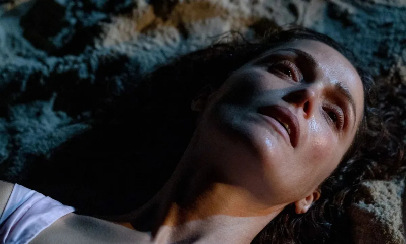 Linda está deitada na areia com uma expressão triste, a câmera está focada em seu rosto.