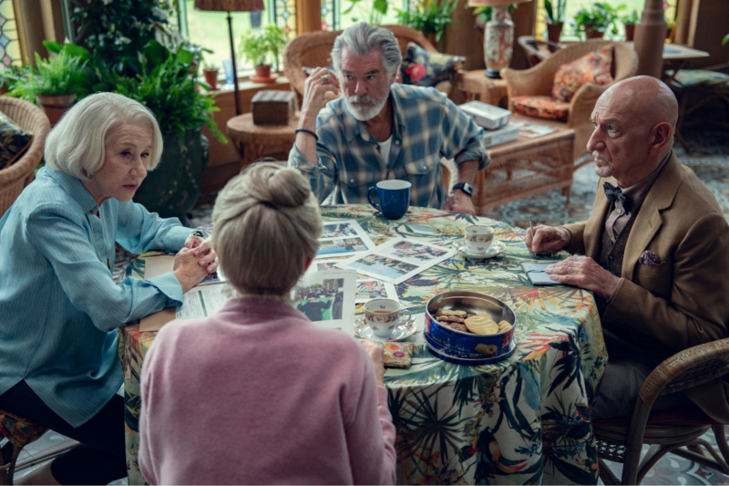 Cena do filme O Clube do Crime das Quintas-Feiras.Na imagem, estão Elizabeth (Helen Mirren), uma mulher branca de cabelos curtos e brancos e cardigan azul, Ron (Pierce Brosnan), um homem branco de cabelos brancos e com camisa xadrez azul e branca, Ibrahim (Ben Kingsley), homem de ascendência indiana e careca vestindo terno e camisa marrons e uma gravata borboleta e Joyce (Celia Imrie), que está de costas, é uma mulher branca de cabelos brancos presos em um coque alto e vestindo um cardigan rosa. Eles estão reunidos em uma sala de estar bem iluminada e decorada com móveis e plantas, sentados ao redor de uma mesa e com expressões de seriedade e atenção. 