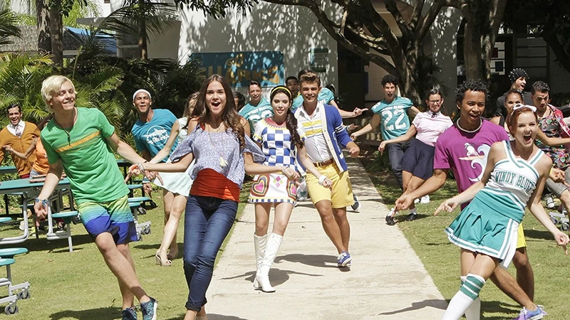 Cena do filme Teen Beach 2. Na imagem, há os personagens Lela, Tanner, Brady e Mack. Da esquerda para direita, a moça branca de cabelos castanhos veste um vestido azul com estampa xadrez azul quadriculada e botas brancas, o homem branco de cabelos castanho claro utiliza uma regata branca com uma blusa vermelha aberta, um short colorido e um tênis branco, o homem branco de cabelos brancos veste uma camiseta laranja e um short azul com estampas de praia e a mulher branca de cabelos castanhos veste uma blusa azul ferida, um short jeans e uma tamanca média marrom. Eles estão em movimento, assim como os figurantes do filme. 