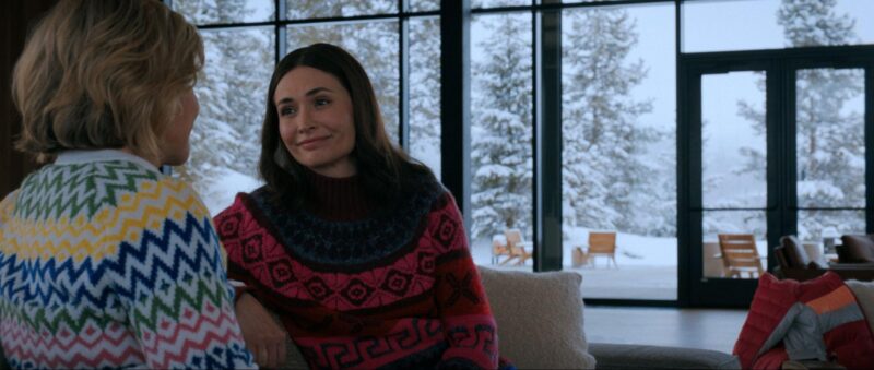 Rhea Seehorn está de costas, posicionada no canto esquerdo da imagem, vestindo um agasalho de fundo branco com traços em verde, amarelo, azul e rosa. À frente, Karolina Wydra, mulher branca de cabelos castanhos e olhos castanhos, está sentada usando um agasalho com traços em rosa e azul. Ao fundo, há um cenário de inverno; as paredes de vidro deixam ver árvores, duas cadeiras e a neve do lado de fora.