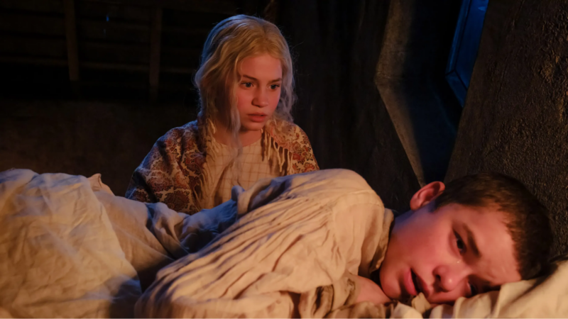 Cena de “O Morro dos Ventos Uivantes”. A versão infantil de Catherine, uma jovem loira observa Heathcliff preocupada. O garoto está deitado de lado na cama, com a face avermelhada, chorando. Ambos vestem roupas simples de época, e o fundo da cena é pouco iluminado.