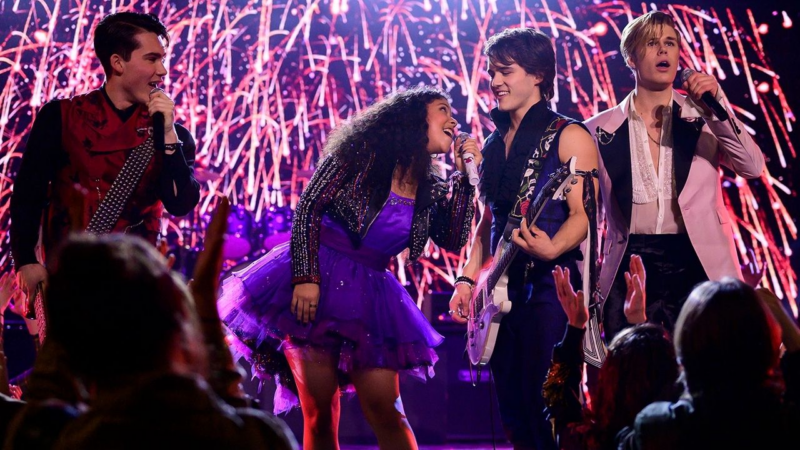 Cena de Julie and the Phantoms. A cena mostra um grupo musical de quatro jovens, composto por três homens e uma mulher, performando em um palco iluminado. A mulher, Julie, é negra e tem cabelos cacheados volumosos, está no centro, vestindo um vestido roxo com saia de tule e jaqueta brilhante. Os homens, brancos, usam roupas estilosas: Reggie com colete vermelho com tachas, Luke com jaqueta azul marinho e sem mangas e Alex com um blazer rosa claro. Todos seguram microfones e estão cantando, com expressões de empolgação. O palco é iluminado por um fundo de luzes vibrantes em tons de vermelho e branco, simulando fogos de artifício. A cena é vista de um ângulo baixo, com a plateia desfocada em primeiro plano, indicando um show ao vivo. A iluminação é intensa e colorida, criando uma atmosfera enérgica e festiva. A composição é dinâmica, com os artistas posicionados de forma a enfatizar a energia da performance.