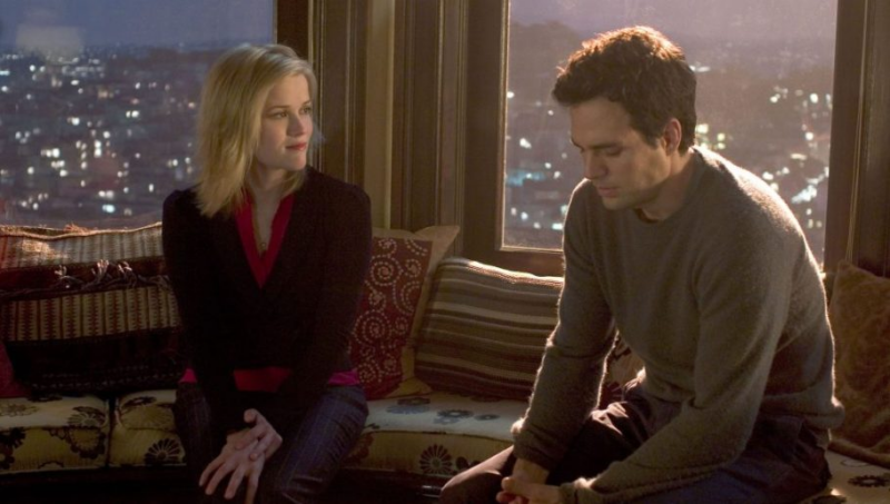 Cena do filme E Se Fosse Verdade. Elizabeth (Reese Witherspoon) e David (Mark Ruffalo) estão sentados em um banco estofado diante de uma ampla janela à noite. Ao fundo, vê-se as luzes desfocadas da cidade de São Francisco. Elizabeth, com um sorriso suave, olha para David, que está sentado com postura curvada, mãos entrelaçadas e olhar baixo, parecendo melancólico