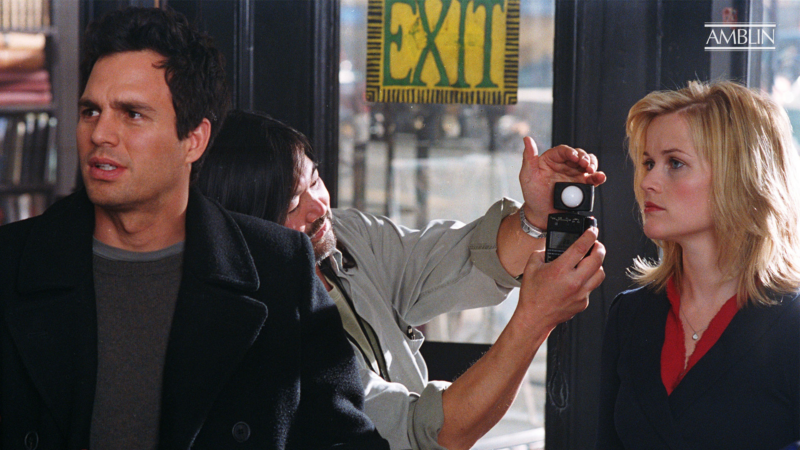 Bastidores do filme E Se Fosse Verdade. Os atores Mark Ruffalo (à esquerda, de casaco preto) e Reese Witherspoon (à direita, de blazer azul marinho) estão parados em uma locação. Entre eles, um membro da equipe de produção segura um fotômetro próximo ao rosto de Reese para medir a luz. Mark olha para o lado com a boca entreaberta, e há uma placa "EXIT" amarela e verde no vidro ao fundo.