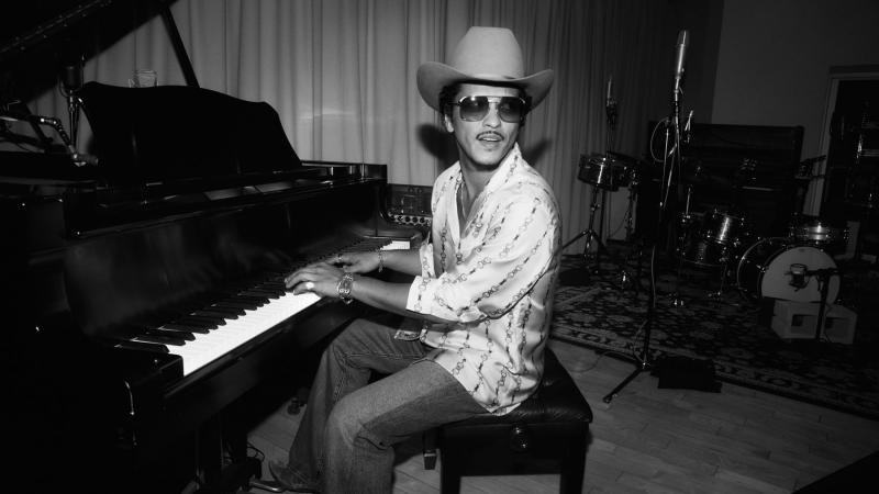 Fotografia de Bruno Mars. Na imagem em preto e branco, Mars aparece tocando um piano em um estúdio. O cantor veste uma camisa listrada, calças escuras, chapéu, pulseiras e óculos escuros. Ele toca o instrumento enquanto se vira e olha para trás. 