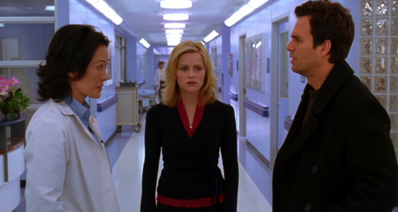 Cena do filme E Se Fosse Verdade. Em um corredor de hospital com paredes e iluminação em tons de azul frio, a Dra. Fran (Rosalind Chao) está à esquerda conversando seriamente com David (Mark Ruffalo), à direita. No centro, Elizabeth (Reese Witherspoon) está parada olhando para frente com uma expressão de preocupação e urgência.