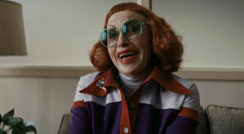 Frame de uma mulher branca de cabelos vermelhos curtos e franja curta sorrindo, como se estivesse no meio de uma risada. Ela usa óculos grandes, quadrados e armação na cor azul, um batom vermelho borrado nos cantos e um casaco listrado lilás, vermelho e azul escuro com uma blusa lilás de gola alta por baixo, sentada em uma sofá cinza. Ao fundo, vemos uma parede bege, lisa, com a parte de baixo revestida em madeira.