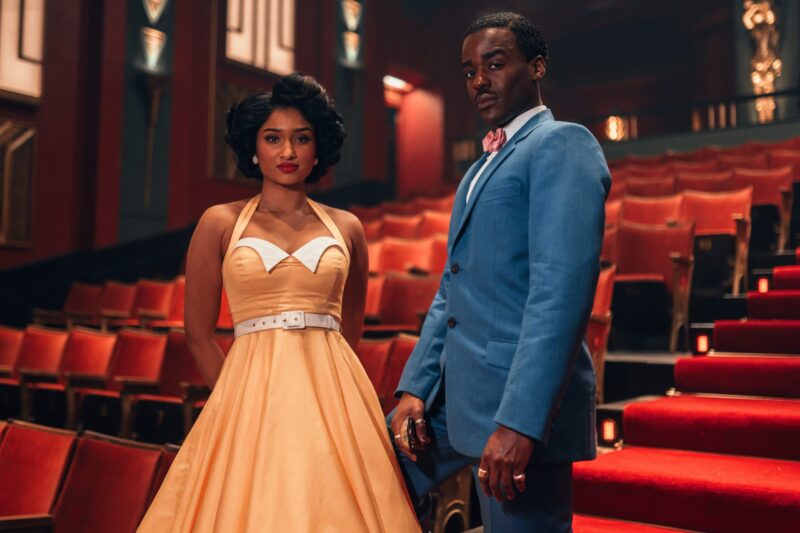 Na imagem, Doutor e Belinda estão em primeiro plano dentro de um cinema antigo com várias fileiras de cadeiras vermelhas. Doutor é um homem negro jovem de cabelo curto escuro e bigode. Ele usa um terno azul por cima de uma camisa branca e uma gravata rosa no colarinho. Belinda está ao lado esquerdo. Ela é uma mulher de pele escura com cabelo moreno penteado no estilo dos anos 60. Ela usa um vestido da mesma época em cor salmão. Ambos os personagens olham diretamente para a câmera.