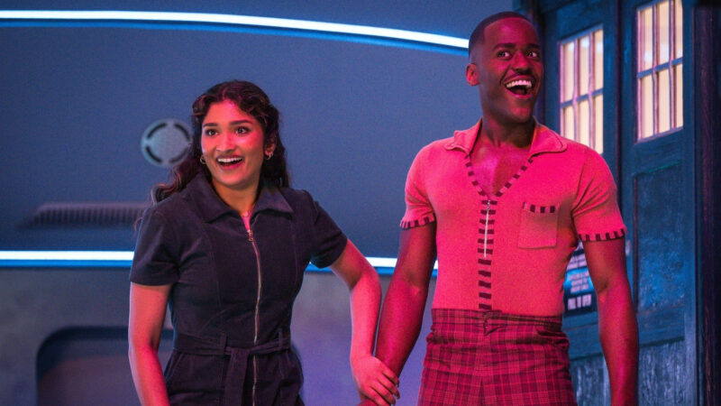 Na imagem, Belinda e Doutor sorriem olhando à esquerda para algo fora de enquadramento. Ela segura o pulso dele. Belinda usa um macacão azul escuro e cabelo enrolado caído sobre os ombros. Doutor está de camiseta laranja de botões e calça xadrez vermelha. Atrás deles há lâmpadas de cores neon futuristas. À direita está a TARDIS, uma cabine telefônica azul com janelas brancas.