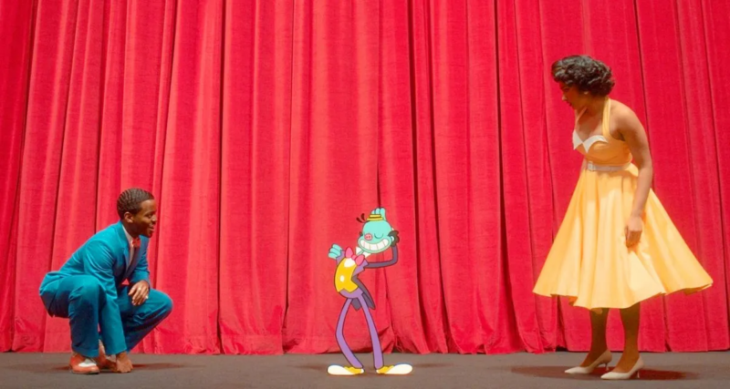 Na imagem, Doutor e Belinda estão em cima de um palco olhando para um personagem animado e pequenino. Doutor está agachado à esquerda, sorrindo, de terno turquesa dos anos 60. Belinda está olhando para baixo de vestido salmão com detalhes brancos dos anos 60 também. O personagem animado tem pele azul, nariz vermelho arredondado e um traje formal roxo e amarelo. O estilo do desenho remete às animações do começo dos anos 30.