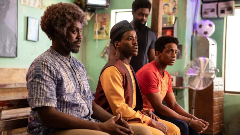 Na imagem, da esquerda para a direita, há 3 homens sentados em um banco e outro em pé atrás deles. Estão em uma barbearia com paredes verdes e quadros diversos ao fundo. O homem mais à esquerda possui pele negra, cabelo crespo grisalho, barba preta e está com as mãos juntas com semblante pensativo. O homem sentado no meio é o Doutor, com roupa amarela típica da Nigéria, colete bronze e um filá escuro na cabeça. Ele está com um semblante assustado. O homem mais à direita possui pele negra, cabelo moreno em topete e camiseta laranja. Por fim, o homem de pé atrás possui pele negra, cabelo e barba morena e está com camiseta preta.