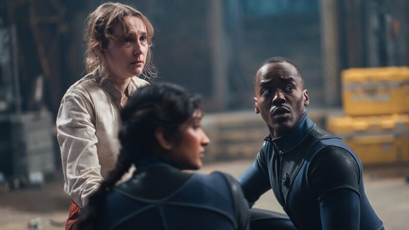 Na imagem, há três pessoas, da esquerda para a direita, a personagem Yeft, Belinda e Doutor. Os três olham para trás com o olhar assustado. Yeft é uma menina branca com cabelo loiro liso. Ela está de camisa branca suja e o cabelo desarrumado. Doutor e Belinda usam trajes espaciais justos na cor azul e com detalhes pretos.