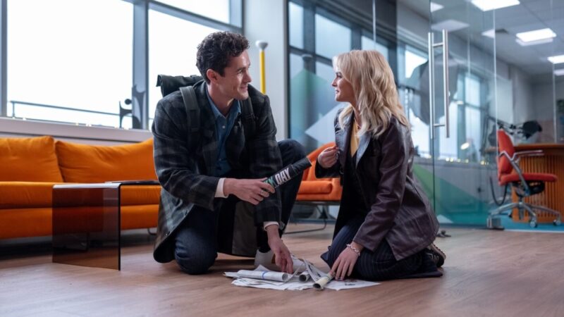 Na imagem, Conrad e Ruby Sunday estão ajoelhados em um escritório moderno, sorrindo um para o outro enquanto organizam papéis espalhados no chão. Ruby é uma mulher branca, loira e jovem. Ela está com roupa social preta, incluindo casaco marrom escuro. Conrad é um homem branco de cabelo moreno e usa roupa social azul com uma jaqueta preta e cinza e mochila quadrada nas costas, o cenário inclui sofá laranja e portas de vidro.