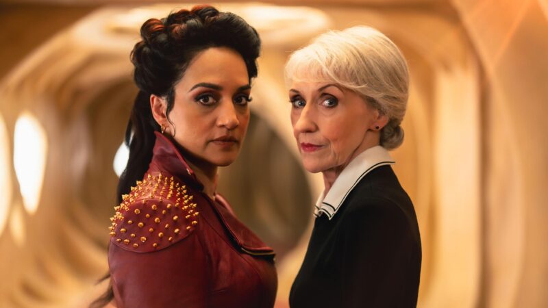 Na imagem, Rani e Mrs. Flood encaram a câmera com expressão séria. Rani é uma mulher de pele parda e cabelos morenos escuros e penteados. Ela veste jaqueta vermelha com ombros cravejados de dourado. Mrs. Flood é uma mulher branca, mais velha, de cabelos brancos e usa roupa formal preta com gola branca. Ambas estão em um corredor futurista iluminado em tons quentes.